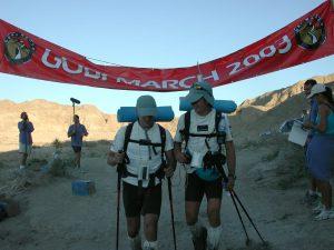 A picture of Mark and his running partner Nick Wolf at the Gobi Desert March, The Race of No Return, 2003. They are in the desert wearing backpacks and gators and peaked caps and are carrying trekking poles