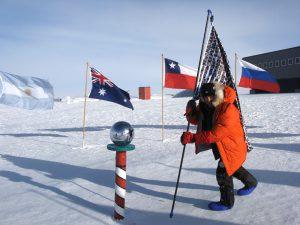 A picture of Mark planting his flag of faces at the South Pole, with other national flags in the background framed by the snow
